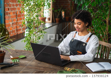 African woman florist gardener working in botanical store using laptop order plants communicating on Internet with customer. Female small business owner working at flower shop. Running of own business 137938124