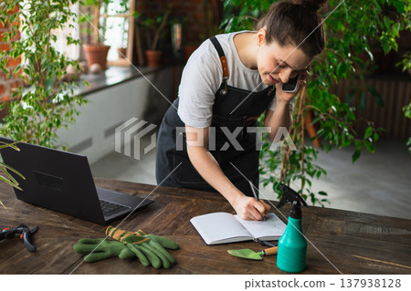 Woman florist gardener working in botanical store greenhouse using laptop order plants by phone communicating with customer. Small business owner working at flower shop. Running of own business Woman florist gardener working in botanical store greenhouse using laptop order plants by phone communicating with customer. Small business owner working at flower shop. Running of own business 137938128