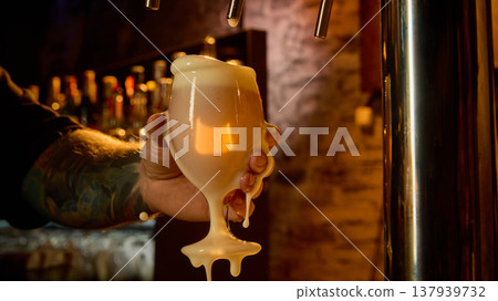 Draft beer overflowing glass with thick foam in bartender hand dark moody bar. Draft beer overflowing glass with thick foam in bartender hand dark moody bar. 137939732