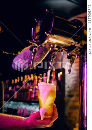 Draft beer pouring into glass with colorful neon lighting and foam overflow in bar. Draft beer pouring into glass with colorful neon lighting and foam overflow in bar. 137939741