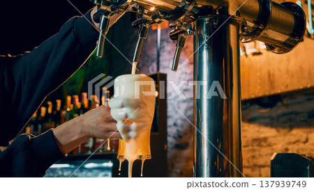 Draft beer overflowing glass in bartender hand with heavy foam in bar 137939749