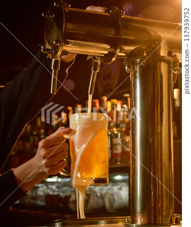 Draft beer pouring into glass mug with foam overflowing in warm bar lighting. 137939752