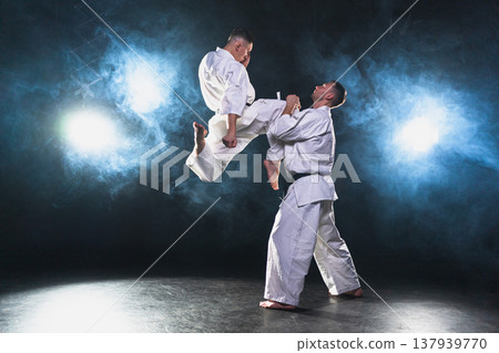 Martial arts masters on dark background. Strong man fighters in white kimono for karate martial arts fighting training in black gym. Karate fighters training punches kicks practicing fight sparring Martial arts masters on dark background. Strong man fighters in white kimono for karate martial arts fighting training in black gym. Karate fighters training punches kicks practicing fight sparring 137939770