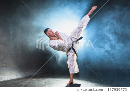 Strong man fighter in white kimono for karate martial arts with black belt preparing for fight training in black background. Man training moves punches kicks practicing shadow fight ready for sparring 137939773