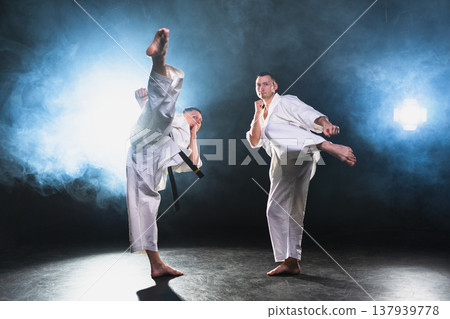 Martial arts masters on dark background. Strong man fighters in white kimono for karate martial arts fighting training in black gym. Karate fighters training punches kicks practicing fight sparring Martial arts masters on dark background. Strong man fighters in white kimono for karate martial arts fighting training in black gym. Karate fighters training punches kicks practicing fight sparring 137939778