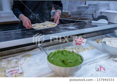 Genoa, liguria, italy. May 18,2024. Hand arranging fresh ravioli beside a large bowl of pesto genovese at an italian market stall, close-up of artisan food display Genoa, liguria, italy. May 18,2024. Hand arranging fresh ravioli beside a large bowl of pesto genovese at an italian market stall, close-up of artisan food display 137940333