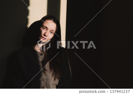 Moody cinematic portrait of a woman or girl in a grey cardigan under dramatic hard sunlight. Authentic emotions in a dark minimalist setting. Moody cinematic portrait of a woman or girl in a grey cardigan under dramatic hard sunlight. Authentic emotions in a dark minimalist setting. 137941237
