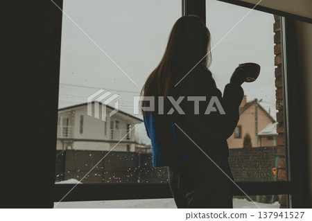 Pensive silhouette of a woman holding a coffee mug, looking out a large window at a snowy suburban winter landscape. Rainy gloomy weather. 137941527