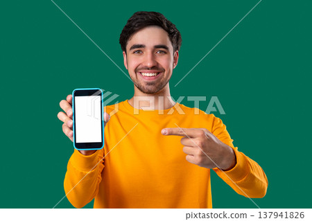 Smiling young man showing smartphone white screen on green background. Mock-up 137941826