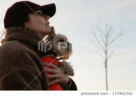 Candid portrait of a woman in a maroon cap and brown fleece jacket holding a small dog in an orange sweater. Lifestyle pet photography, emotional bond Candid portrait of a woman in a maroon cap and brown fleece jacket holding a small dog in an orange sweater. Lifestyle pet photography, emotional bond 137942391