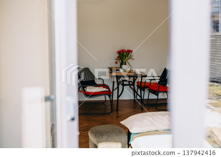 Cozy studio apartment interior with a dining table, red chairs, and fresh tulips. Modern minimalist decor, natural light, lifestyle home atmosphere Cozy studio apartment interior with a dining table, red chairs, and fresh tulips. Modern minimalist decor, natural light, lifestyle home atmosphere 137942416