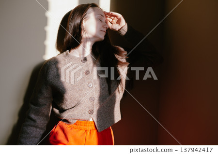 Serene young white woman basking in warm sunlight indoors, wearing cozy brown cardigan. Mindfulness, slow living, home atmosphere, mental wellness Serene young white woman basking in warm sunlight indoors, wearing cozy brown cardigan. Mindfulness, slow living, home atmosphere, mental wellness 137942417