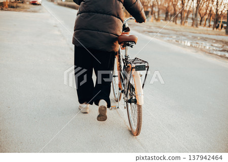 Back view of a woman walking with a vintage brown bicycle on a city or suburb road. Autumn or spring season, urban transportation. Lifestyle scene. Back view of a woman walking with a vintage brown bicycle on a city or suburb road. Autumn or spring season, urban transportation. Lifestyle scene. 137942464
