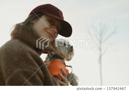 Candid portrait of a woman in a maroon cap and brown fleece jacket holding a small dog in an orange sweater. Lifestyle pet photography, emotional bond Candid portrait of a woman in a maroon cap and brown fleece jacket holding a small dog in an orange sweater. Lifestyle pet photography, emotional bond 137942467