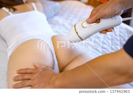 Women getting help with VIO hair removal from their partners (boyfriends, husbands) while lying face down; using home hair removal devices; trimming up. Women getting help with VIO hair removal from their partners (boyfriends, husbands) while lying face down; using home hair removal devices; trimming up. 137942963