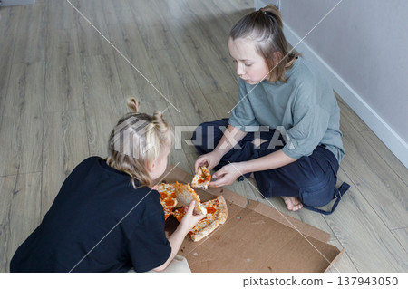 Lovely young children sitting on floor in living room and eating pizza at home Lovely young children sitting on floor in living room and eating pizza at home 137943050