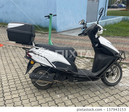 White scooter parked near a blue building on a paved area with green space in the background  137943153