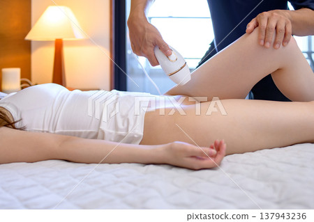 A woman gets help with VIO hair removal from her partner (boyfriend, husband) using a home hair removal device; side view. A woman gets help with VIO hair removal from her partner (boyfriend, husband) using a home hair removal device; side view. 137943236