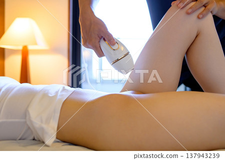 A woman gets help with VIO hair removal from her partner (boyfriend, husband) using a home hair removal device; side view. A woman gets help with VIO hair removal from her partner (boyfriend, husband) using a home hair removal device; side view. 137943239