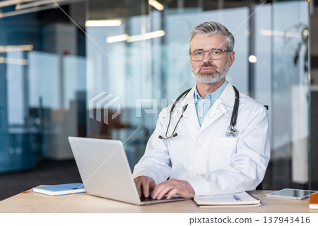 Senior male doctor with eyeglasses and stethoscope sitting at his desk, typing on a laptop, providing professional medical consultation and healthcare services in a clinic environment Senior male doctor with eyeglasses and stethoscope sitting at his desk, typing on a laptop, providing professional medical consultation and healthcare services in a clinic environment 137943416