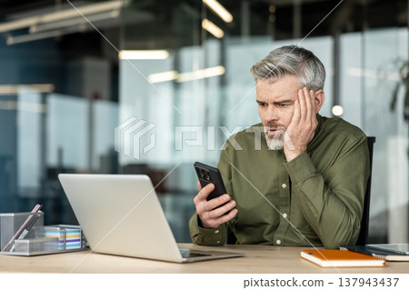 Mature man sitting at an office desk, looking distressed while checking his mobile phone, experiencing stress or anxiety from a difficult message or result Mature man sitting at an office desk, looking distressed while checking his mobile phone, experiencing stress or anxiety from a difficult message or result 137943437