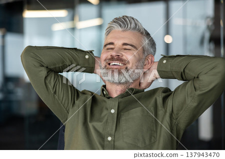 Mature man with grey hair and beard leaning back with hands behind his head, smiling with eyes closed, embodying business success, relaxation, and satisfaction in a modern office environment 137943470