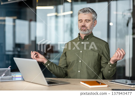 Mature man finding zen, meditating with closed eyes and mudra gesture at his desk with a laptop, practicing mindfulness to relieve stress and boost focus in a modern office environment 137943477