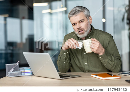 Mature man sitting at his office desk, holding a tissue and a mug, experiencing symptoms of a cold or flu while trying to continue working, highlighting workplace presenteeism 137943480