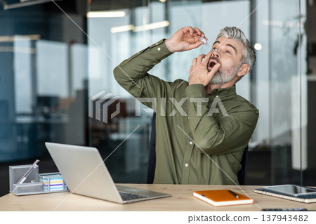 Mature businessman applying moisturizing eye drops to alleviate dry eyes and computer vision syndrome caused by prolonged screen time while working in modern office environment 137943482