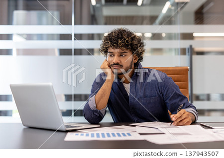 Young Indian businessman sitting at his desk in the office, looking bored and tired at his laptop. 137943507