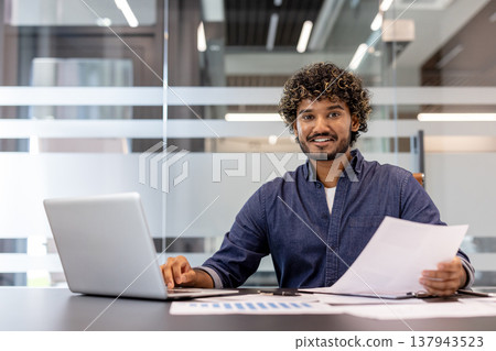 Portrait of a smiling and successful Indian businessman sitting in the office at a table with a laptop, holding documents in his hands and looking at the camera. Portrait of a smiling and successful Indian businessman sitting in the office at a table with a laptop, holding documents in his hands and looking at the camera. 137943523