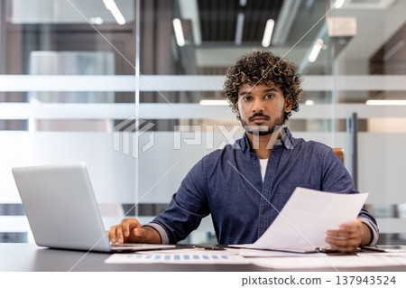 Portrait of a young Indian man sitting at a desk in the office, working with a laptop and documents. 137943524