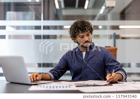 A serious young Indian man is working intently in the office on a laptop and making notes in documents. A serious young Indian man is working intently in the office on a laptop and making notes in documents. 137943540