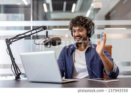 Smiling young Indian man in headphones sitting in the office at a table with a laptop and talking on a microphone, greeting and waving at the camera. Smiling young Indian man in headphones sitting in the office at a table with a laptop and talking on a microphone, greeting and waving at the camera. 137943550
