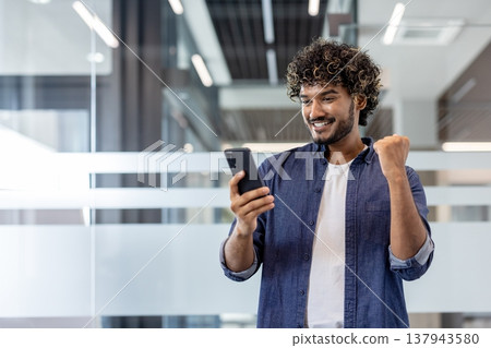 An Indian man standing in the office, looking at the phone screen and rejoicing in success and winnings. 137943580