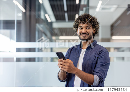 Portrait of a young Indian man standing in an office, holding a mobile phone and looking at the camera with a smile. 137943584