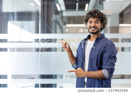 Portrait of a smiling Indian young man standing in the office and pointing with his fingers at an empty space. Portrait of a smiling Indian young man standing in the office and pointing with his fingers at an empty space. 137943594