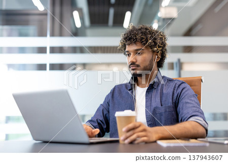 Serious and focused young Indian man working with headphones on a laptop in the office, sitting in a chair, drinking coffee. 137943607