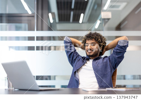 Happy young Indian man is reading in the office at a table in front of a laptop, hands behind his head and smiling. Happy young Indian man is reading in the office at a table in front of a laptop, hands behind his head and smiling. 137943747