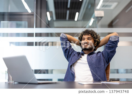 A young Indian man is satisfied with the final result, sitting in an office chair, hands behind his head, and resting. A young Indian man is satisfied with the final result, sitting in an office chair, hands behind his head, and resting. 137943748