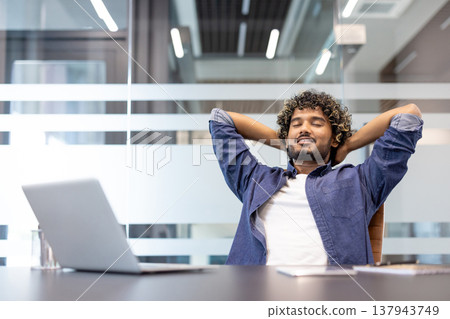 Relaxed young Indian man with his hands behind his head and eyes closed resting in a chair in the office. Relaxed young Indian man with his hands behind his head and eyes closed resting in a chair in the office. 137943749