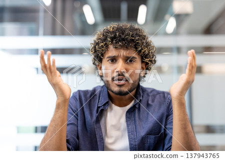 Close-up photo of a worried young Indian man sitting at a desk in an office facing the camera and talking emotionally. Close-up photo of a worried young Indian man sitting at a desk in an office facing the camera and talking emotionally. 137943765