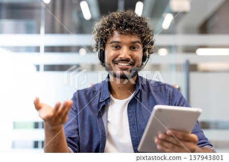 Portrait of a young smiling Indian man in a headset sitting at a desk in the office in front of the camera and talking on a video call. 137943801
