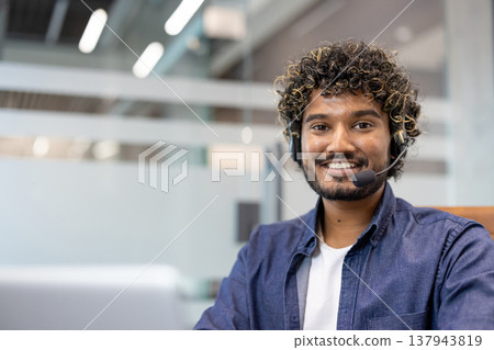 Close-up portrait of a young Indian man in a headset working in an office on a laptop and looking at the camera with a smile. Close-up portrait of a young Indian man in a headset working in an office on a laptop and looking at the camera with a smile. 137943819