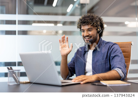 Young Indian man in headset sitting at a desk in the office, talking on a video call on a laptop, greeting and waving. Young Indian man in headset sitting at a desk in the office, talking on a video call on a laptop, greeting and waving. 137943824