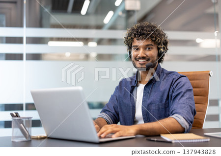 Portrait of a smiling young Muslim man in a headset working in an office at a laptop and looking at the camera. Portrait of a smiling young Muslim man in a headset working in an office at a laptop and looking at the camera. 137943828