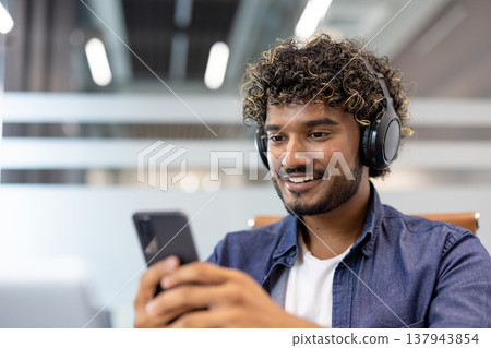 Close-up photo of an Indian young man in an office wearing headphones and using a phone. 137943854