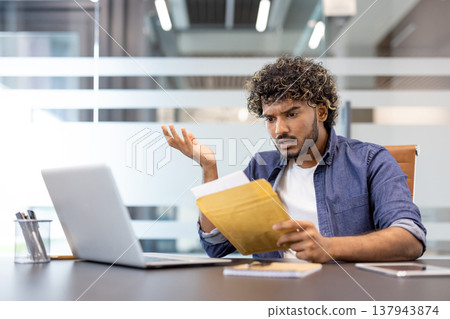 A worried young Indian man sits at his desk in the office and looks disappointedly at the letter he received. A worried young Indian man sits at his desk in the office and looks disappointedly at the letter he received. 137943874
