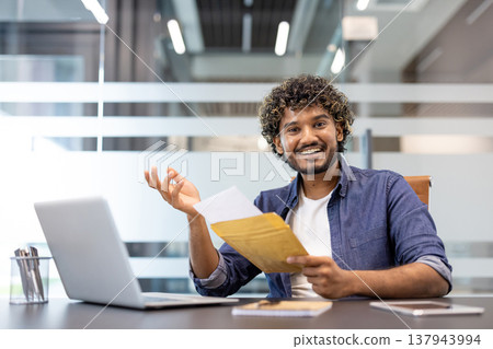 Portrait of a young Muslim man sitting in an office at a desk, holding an envelope with a letter and smiling at the camera. Portrait of a young Muslim man sitting in an office at a desk, holding an envelope with a letter and smiling at the camera. 137943994