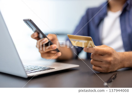 A close-up photo of the hands of a young man in an office, sitting in front of a laptop, holding a credit card and a phone. A close-up photo of the hands of a young man in an office, sitting in front of a laptop, holding a credit card and a phone. 137944008